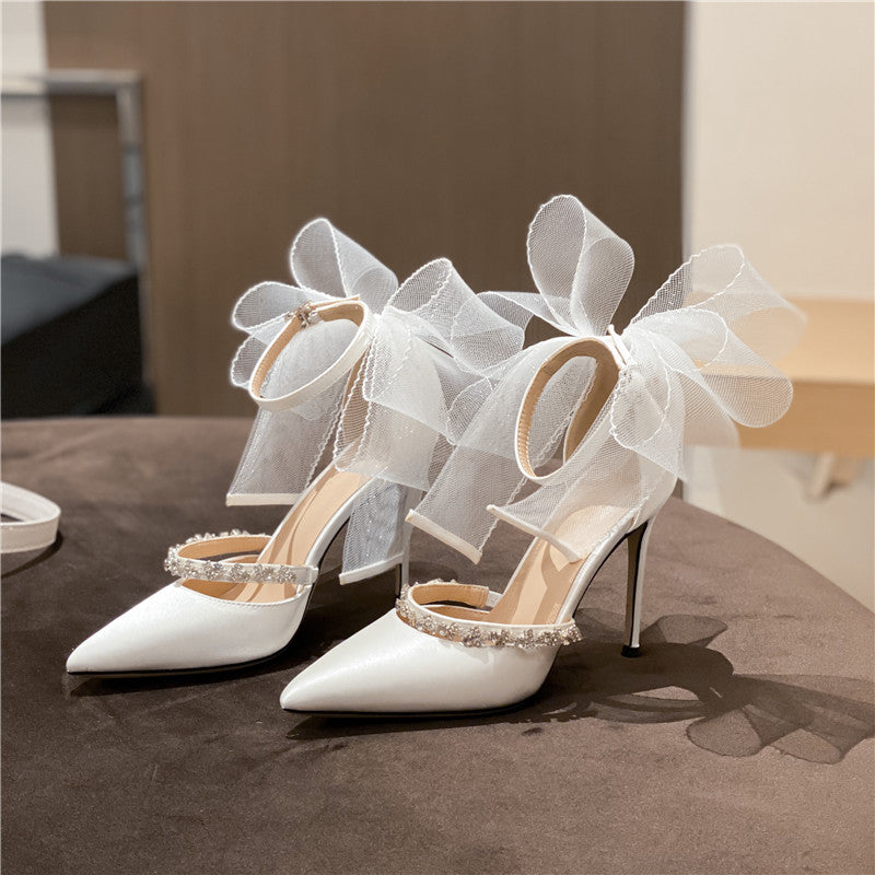 White high-heeled wedding sandals with rhinestone bowknot and ankle strap, pointed toe design, displayed on a brown surface.