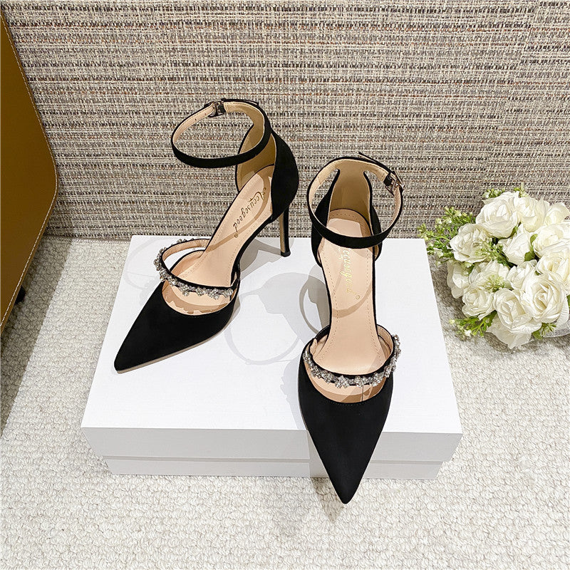 Black high-heeled sandals with rhinestone bow detailing, pointed toe, and ankle strap on display with white roses in the background.