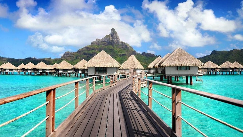 Overwater bungalows on turquoise lagoon with mountain backdrop under blue sky and white clouds